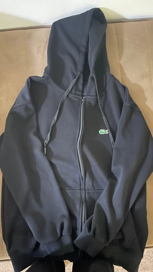 Svart hoodie från Lacoste med dragkedja - Svart hoodie från Lacoste med klassisk krokodillogga på bröstet. Tröjan har huva med snörning, hel dragkedja framtill och ribbade muddar. Regular fit och tillverkad i mjuk bomullsmix. Perfekt för chill dagar eller när du vill ha en clean streetstyle.