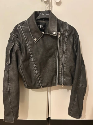 Svart croppad skinnjacka från Zara - Säljer en svart croppad skinnjacka från Zara med coola detaljer som snedställda dragkedjor och silvriga knappar. Jackan har klassisk krage och lång ärm, perfekt för dig som vill ha en edgy look. Passformen är normal och materialet är syntetiskt skinn.