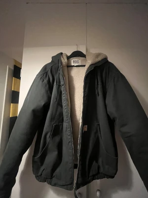 Mörkgrön vinterjacka från BDG Urban Outfitters - Mörkgrön teddyjacka från BDG Urban Outfitters med mjukt beige foder och huva. Jackan har dragkedja framtill, två stora fickor och elastisk nederkant. Utsidan är i slitstark bomull och insidan i mysig fleece. Perfekt för kalla dagar och chill stil.