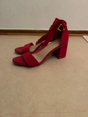 Röda klackskor Säljes!  - Snygga röda klackskor från Deichmann med breda remmar och chunky klack. Skorna har öppen tå och spänne i guld vid ankeln. Tillverkade i mocka-look som ger en mjuk och lyxig känsla. Använd bara max två gånger 