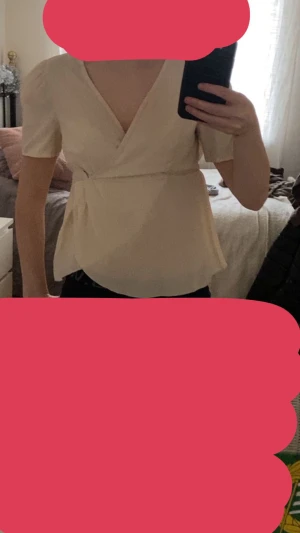 Beige omlottblus med v-ringning - Säljer en beige omlottblus med korta puffärmar och v-ringad hals. Blusen har en markerad midja och ett lätt utsvängt fall nedtill. Materialet är mjukt och har en lätt glans, vilket ger en fräsch och stilren look. Perfekt för dig som gillar enkla men snygga detaljer.