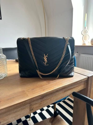 Svart quiltad axelväska från Saint Laurent - Säljer en svart quiltad axelväska från Saint Laurent med guldiga kedjor och YSL-logga framtill. Väskan är tillverkad i skinn och har ett elegant mönster med V-formade sömmar. Perfekt storlek för att få plats med allt du behöver och ger en lyxig vibe till din outfit.