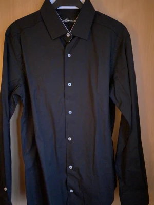 Svart skjorta från Amacci - Stilren svart skjorta från Amacci med klassisk krage och vita knappar framtill. Skjortan har lång ärm och en rak passform som funkar till många olika looks. Materialet känns lätt och bekvämt mot huden. Perfekt för dig som gillar enkel och snygg stil.