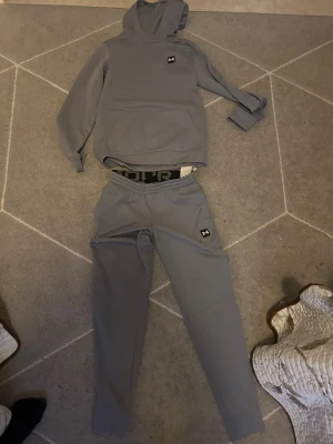 Grå tracksuit från Under Armour - Säljer en stilren grå tracksuit  från Under Armour med klassisk logga på bröstet. Tröjan har huva, lång ärm och en stor magficka framtill. Materialet är mjukt och känns sportigt, perfekt för chill eller träning. Snyggt tryck med Under Armour på baksidan av midjan.