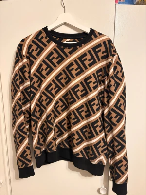 Fendi brun och svart sweatshirt L - Säljer en fet Fendi sweatshirt i storlek L med det klassiska FF-mönstret i svart och brunt. Tröjan har rund hals, ribbade muddar och är långärmad. Materialet är mjuk bomull med insida i fleece, perfekt för dig som gillar lyxiga streetwear vibes.