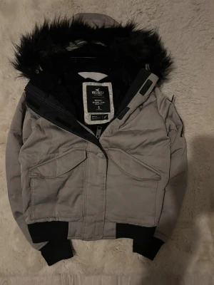 Grå dunjacka med svart fuskpäls Hollister - Säljer en grå dunjacka från Hollister Ultimate Down Collection i storlek S. Jackan har svart fuskpäls på huvan, stora fickor framtill, ribbade muddar och dragkedja. Fylld med dun och fjäder för extra värme, perfekt för kalla dagar.