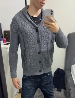 Grå stickad kofta med knappar - Säljer en grå stickad kofta med grovt kabelmönster framtill och bred krage. Koftan har fem stora svarta knappar och ribbade muddar vid ärmslut och nederkant. Perfekt för dig som gillar chill och stilren look.