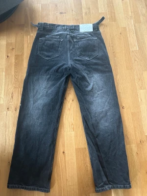 Svarta vicinity jeans med tvättat look - Säljer ett par svarta jeans med tvättad look och raka ben. Jeansen har fem fickor, bälteshällor och klassisk knappgylf. Passformen är loose och benen är breda hela vägen ner. Perfekta för dig som gillar avslappnad stil.