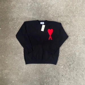 Svart stickad tröja från Ami - Svart stickad tröja från Ami med rund hals och lång ärm. På bröstet finns ett stort rött hjärta och bokstaven A som ger en unik och snygg detalj. Tröjan har ribbade muddar vid ärmslut och nederkant för extra komfort.