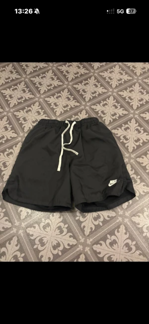 Svarta Nike shorts med snörning - Svarta shorts från Nike i storlek S, med vit snörning i midjan och klassisk Nike-logga på benet. De har elastisk midja, bakficka med lock och är gjorda i lätt polyester med meshfoder. Perfekta för träning eller chill dagar.