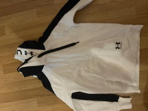 Vit och svart hoodie från Under Armour - Säljer en snygg vit och svart hoodie från Under Armour med stor logga på huvan och bröstet. Tröjan har lång ärm, huva med snörning och en stor magficka. Perfekt för träning eller chill. Materialet är mjuk bomull och polyester.