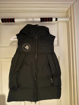 Svart dunväst från Sail Racing - Svart dunväst från Sail Racing i storlek XS med huva och tvåvägsdragkedja. Använd fåtal gånger.    Nypris: 3500kr