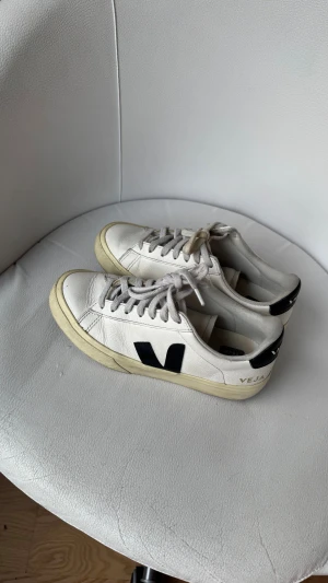 Vita Veja sneakers med svart detalj - Säljer ett par klassiska Veja sneakers i vitt skinn med svarta detaljer och logga på sidan och hälen. Skorna har rund tå, platt sula och snörning. Perfekta för en clean och stilren look.