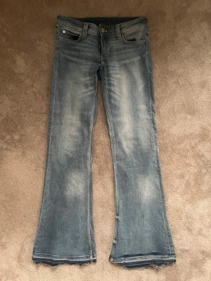 Ljus blå bootcut jeans från Cheap Monday - Säljer ett par ljusblå bootcut jeans från Cheap Monday med råa, fransiga benslut och klassisk femficksdesign. Jeansen har en snygg tvättad look och ett stort grafiskt märke bak i midjan. Perfekta för dig som gillar retrovibbar och streetstyle.