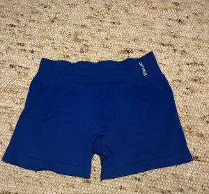 Blå träningsshorts från DFYNE - Säljer ett par blå seamless träningstights-shorts från DFYNE. De har hög midja och är gjorda i ett stretchigt syntetmaterial som sitter tight och bekvämt. Perfekta för gymmet eller löpning. Loggan syns framtill och baktill i vitt. Lappen på insidan är bortskavd men det är en medium