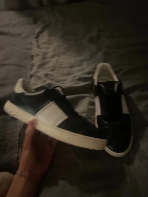 Svarta och vita Valentino flycrew sneakers - Säljer ett par snygga Valentino Open sneakers i svart och vitt med klassisk bred vit rem över sidan. Skorna har platt sula, rund tå och coola nitar på hälen. Perfekta för dig som vill ha en stilren men lyxig look. Storlek 43 passar 44