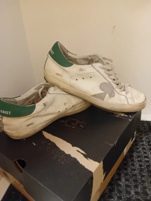 Vit sneaker med gröna detaljer - Säljer ett par vita sneakers från The Galerist med gröna detaljer på hälen och ett stort klubbmärke på sidan. Skorna har snörning, platt sula och rund tå. Materialet är skinn och syntet, med svart mönster på hälen. Perfekt för dig som gillar unik design.Med lite lim så kan dom bli i riktigt bra skick. Ny priss 2100 mitt priss 350