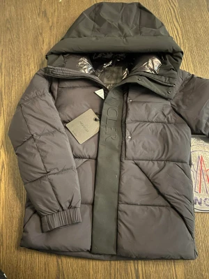Moncler madeira jacka  - Säljer en svart pufferjacka från Moncler med stor huva och tydlig logga på bröstet. Jackan har quiltad design, dragkedja och tryckknappar framtill samt elastiska muddar vid ärmsluten. Perfekt för kalla dagar och riktigt snygg streetstil. Helt ny 