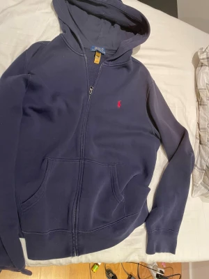 Mörkblå hoodie från Polo Ralph Lauren - Snygg mörkblå hoodie från Polo Ralph Lauren med dragkedja och huva. Tröjan har ett rött broderat Polo-logo på bröstet, två fickor framtill och är tillverkad i mjuk bomull. Perfekt för chill dagar och enkel att matcha med jeans eller joggers den är igentligen 18-20 på barn men sitter som S.  