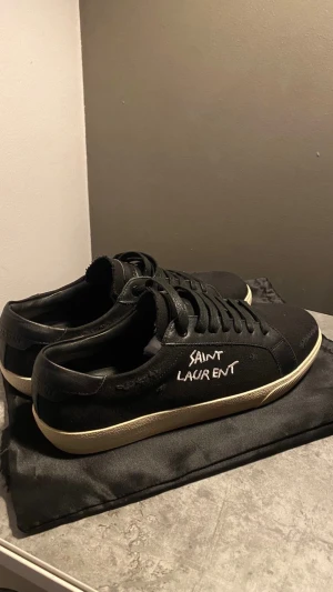 Svarta Saint Laurent sneakers - Svarta sneakers från Saint Laurent med vit handskriven logga på sidan. Skorna har en rund tå, snörning och en kontrasterande beige platt sula med fiskbensmönster undertill. Ovandelen är i canvas och skinn, vilket ger en cool och stilren vibe.