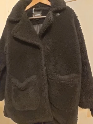 Svart teddyjacka från Monki - Mysig svart teddyjacka från Monki med stora framfickor och knappar framtill. Jackan har en oversized passform, bred krage och är helfodrad för extra värme. Perfekt för kalla dagar när du vill vara både varm och snygg.
