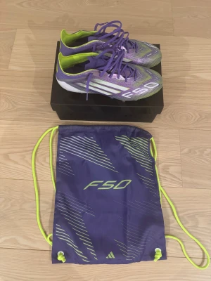 Adidas F50 Elite AG fotbollsskor 42 2/3 - Adidas F50 Elite AG fotbollsskor i lila och neon-gult med vita detaljer. Skorna har snörning, platt sula med dobbar för konstgräs och ett sportigt, randigt mönster. Ovandelen är tillverkad i syntetmaterial och kommer med en matchande F50-påse.