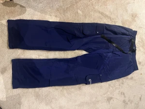 Mörkblå tracksuit byxor från Sergio Tacchini - Säljer ett par mörkblå tracksuit byxor från Sergio Tacchini med raka ben och elastisk midja. Byxorna har flera praktiska fickor med lock och knapp, samt ett justerbart bälte i midjan. Materialet är lätt och syntetiskt, perfekt för chill och streetwear.