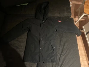 Svart parkas från Canada Goose S - Säljer en svart parkas från Canada Goose med huva och klassisk logga på ärmen. Jackan har knäppning med stora knappar framtill och två snedställda fickor. Perfekt för kalla dagar och riktigt snygg är i bra skick 
