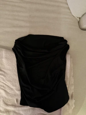 Svart ärmlös tröja - Snygg svart kortklänning med ärmlös design och draperad front. Klänningen har en stilren och enkel look som passar perfekt för dig som gillar minimalistisk stil. Materialet känns mjukt och har en lätt glansig finish som ger en exklusiv känsla.