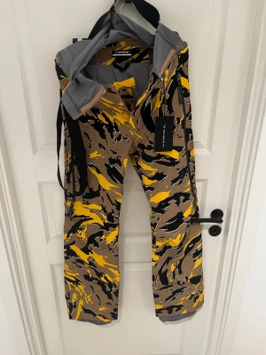 J.Lindeberg camo snowboardbyxor S - Säljer ett par täckbyxor från J.Lindeberg i storlek S med grymt camouflagemönster i brunt, gult och svart. Byxorna har justerbara hängslen, dragkedjor i sidan och är tillverkade i slitstarkt syntetmaterial. Perfekta för vinterns äventyr och riktigt snygga detaljer. Helt nya med prislapp kvar! Skidbyxor eller snowboardbyxor