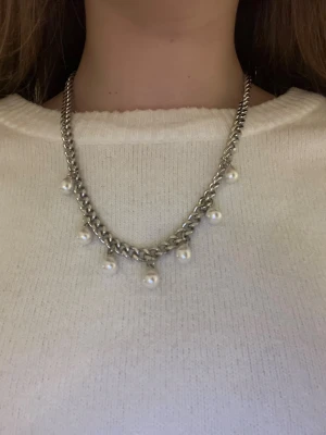 Silverfärgat halsband med pärlor - Stilrent halsband i silverfärgad metall med grov kedja och hängande vita pärlor. Perfekt för att ge din outfit en cool och elegant touch. Kedjan har en chunky känsla och pärlorna ger en snygg kontrast mot metallen. Rostfritt stål och handgjort 