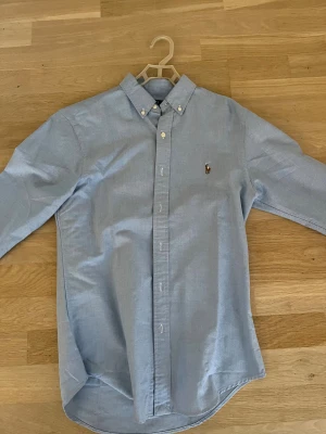 Ljusblå skjorta Polo Ralph Lauren - Klassisk ljusblå skjorta från Polo Ralph Lauren i slim fit. Skjortan har button-down krage, broderad logga på bröstet och är tillverkad i mjuk bomull. Perfekt för en clean och stilren look. Långärmad och med knappar hela vägen.