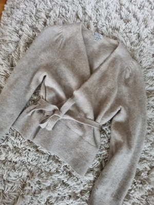 Beige omlottkofta från & Other Stories - Supermysig beige omlottkofta från & Other Stories Paris Atelier. Koftan har knytband i midjan, lång ärm och är stickad i ett mjukt material. Perfekt för lager-på-lager och chill dagar.