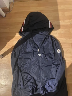 Mörkblå vindjacka från Moncler - Snygg mörkblå vindjacka från Moncler med huva och coola vita och röda ränder på sidan av huvan. Jackan har dragkedja framtill, snörning vid huvan och Moncler-logga på ärmen. Tillverkad i lätt polyester som är perfekt för blåsiga dagar. Helt ny 