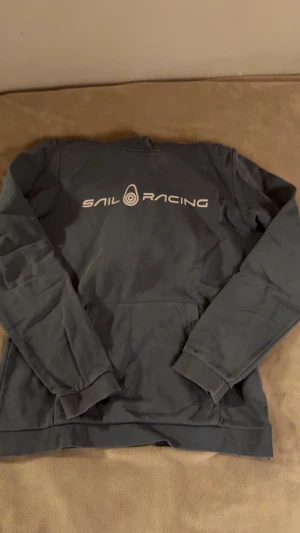 Mörkblå Sail Racing hoodie - Snygg mörkblå hoodie från Sail Racing med stort vitt logotryck på bröstet. Tröjan har lång ärm, klassisk känguruficka och ribbade muddar. Perfekt för dig som gillar sportig och avslappnad stil.
