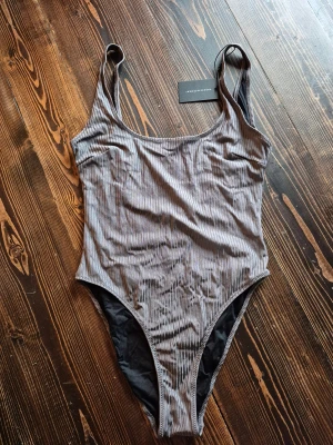 Grå ribbad baddräkt från Understatement - Supersnygg grå baddräkt från Understatement i storlek M. Den har ett stilrent randigt mönster och är tillverkad i ett mjukt syntetmaterial som sitter skönt mot kroppen. Modellen har breda axelband och en klassisk, något hög benskärning och öppen i ryggen. Perfekt för strandhäng eller poolparty!