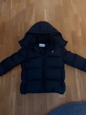 Svart pufferjacka från Calvin Klein Jeans - Svart pufferjacka från Calvin Klein Jeans med huva och dragkedja framtill. Jackan har logga på bröstet och ärmen, samt två sidofickor med dragkedja. Perfekt för kalla dagar och har en clean, modern look, jackan är i storlek M men passar som S, nypris på jackan är 2799kr, jag säljer den för 2000 men priset kan prutas prutas 💨 jackan är väldigt varm och skön, och passar perfekt nu till vintern. 🥰