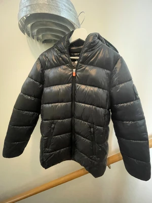 Svart pufferjacka från Save The Duck - Säljer en svart pufferjacka från Save The Duck med huva och dragkedja framtill. Jackan är quiltad och tillverkad i 100% animal free syntetmaterial. Två fickor med dragkedja på sidorna. Perfekt för kalla dagar och riktigt snygg streetstil.