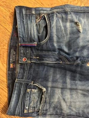 Blå Replay FC Barcelona jeans Anbass  - Snygga blå jeans från Replay i samarbete med FC Barcelona. Anbass modell Modellen har slitna detaljer, smal passform och klassisk femficksdesign. Jeansen har röda och blå detaljer vid knapp och hällor samt patch bak. Perfekt för dig som gillar sportig streetstyle. Obs Dom är lagade, se bilder.