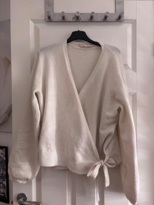 Ljusbeige/vit omlottkofta Pernilla Wahlgren - Supermjuk ljusbeige/vit omlottkofta från Pernilla Wahlgren med knyt i midjan och ribbade muddar. Koftan har lång ärm och en avslappnad passform, perfekt för lager-på-lager. Stilren och enkel att matcha med allt.