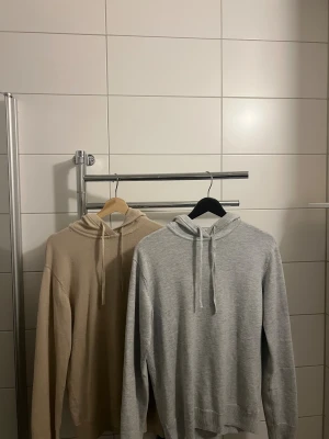 Beige och grå hoodie från RYVLS - Två stilrena hoodies från RYVLS Stockholm, en i beige och en i ljusgrått. Båda har huva med snörning och är ribbstickade vid ärmslut och nederkant. Perfekt för dig som gillar enkel och clean stil.