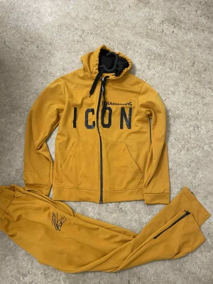 Mustardgul hoodie DSQUARED2 ICON - Säljer en snygg hoodie i mustardgul färg från DSQUARED2 med 'ICON' och 'RAHIMOVIC' tryck på framsidan. Baksidan har ett stort grafiskt tryck och text. Hoodien har huva med snörning, dragkedja och långa ärmar. Materialet är mjuk bomull, perfekt för chill dagar.