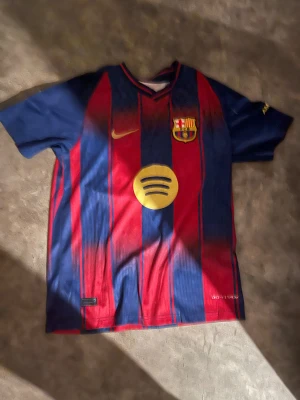 FC Barcelona matchtröja Nike röd/blå - Säljer en FC Barcelona matchtröja från Nike med klassiska röda och blå vertikala ränder. Tröjan har klubbmärket, gul Nike-logga och Spotify som sponsor framtill, samt UNHCR-logga på ryggen. Kortärmad och tillverkad i lätt polyester för maximal komfort på planen.