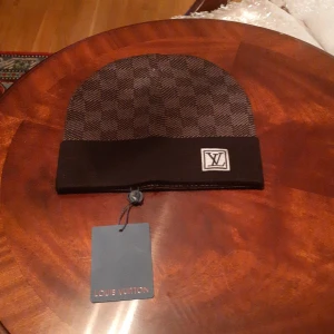 Louis Vuitton rutig brun mössa - Exklusiv mössa från Louis Vuitton i brunt med klassiskt rutigt mönster och bred uppvikt kant. Märkt med LV-logga framtill och etikett på insidan. Materialet är stickat och känns lyxigt och mjukt, perfekt för dig som vill ha en snygg accessoar med designer-vibe.