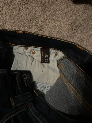 Mörkblå jeans från Nudie Jeans, 28/32 - Snygga mörkblå jeans från Nudie Jeans i klassisk femficksmodell. Jeansen har kontrastsömmar i orange och en rak passform som sitter schysst på benen. Materialet är slitstarkt denim och färgen är djupblå med lätt tvättad look. Perfekt för dig som gillar stilrena jeans.