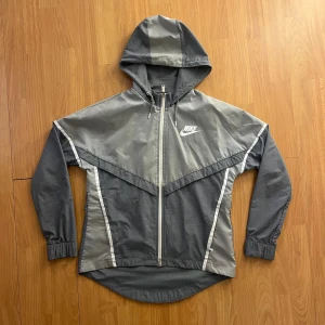 Grå Nike vindjacka med huva - Snygg grå vindjacka från Nike med vit logga på bröstet och dragkedja framtill. Jackan har huva, långa ärmar med resår vid handlederna och coola kontrastpaneler i olika grå nyanser. Perfekt för dig som gillar sportig stil och vill ha något lätt och funktionellt.