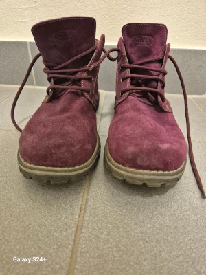 Vinröda mockakängor från Kavle - Säljer ett par vinröda kängor från Kavle i mjuk mocka med grov sula och snörning. Skorna har rund tå och kontrastsömmar, samt brunt foder och plös i skinn. Perfekta för dig som vill sticka ut med färg i höst och vinter.