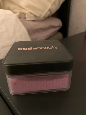 Huda Beauty Easy Bake Ube Birthday Cake - Lila löspuder från Huda Beauty i nyansen Ube Birthday Cake. Kommer i en fyrkantig plastburk med svart lock och roséguldfärgad logga. Perfekt för baking och att fixera sminket med en matt finish. Innehåller 20g.