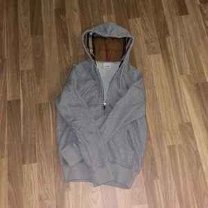 Burberry zip grå - Grå hoodie från Burberry med design i luvan. Tröjan har snörning i huvan, två fickor och ribbade muddar. Storlek XS, midja 43cm, längd 58cm.