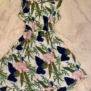 Blommig sommarklänning med resårmidja - Superfin kort klänning med tropiskt blommönster i grönt, blått och rosa på vit botten. Klänningen har ärmlös design, rund halsringning och resår i midjan för en skön passform. Perfekt för varma dagar och ger en fräsch vibe.